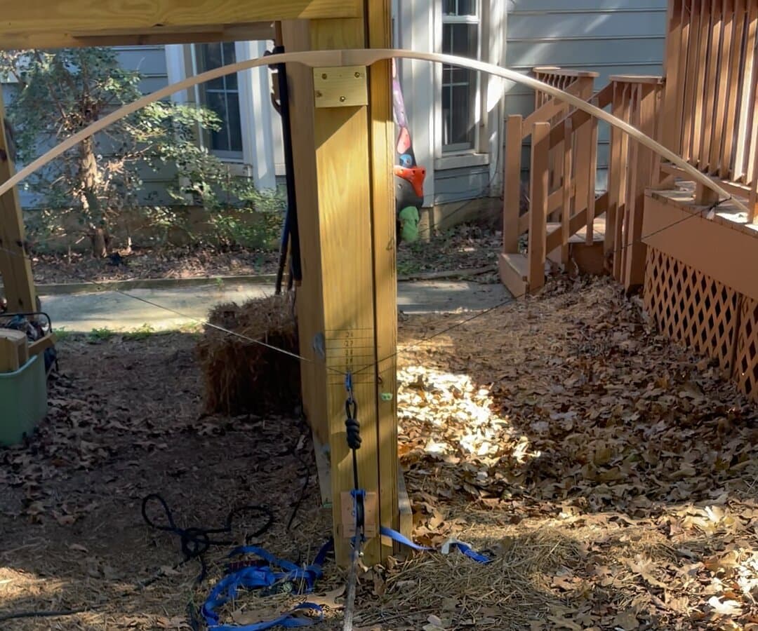 A wooden bow braced at its grip on a wooden post, bent by a string pulled down toward the camera